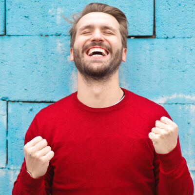 homme de face avec un pull rouge qui semble satisfait, les yeux fermés la tête légèrement penchée en arrière, un grand sourire et les poings serrés