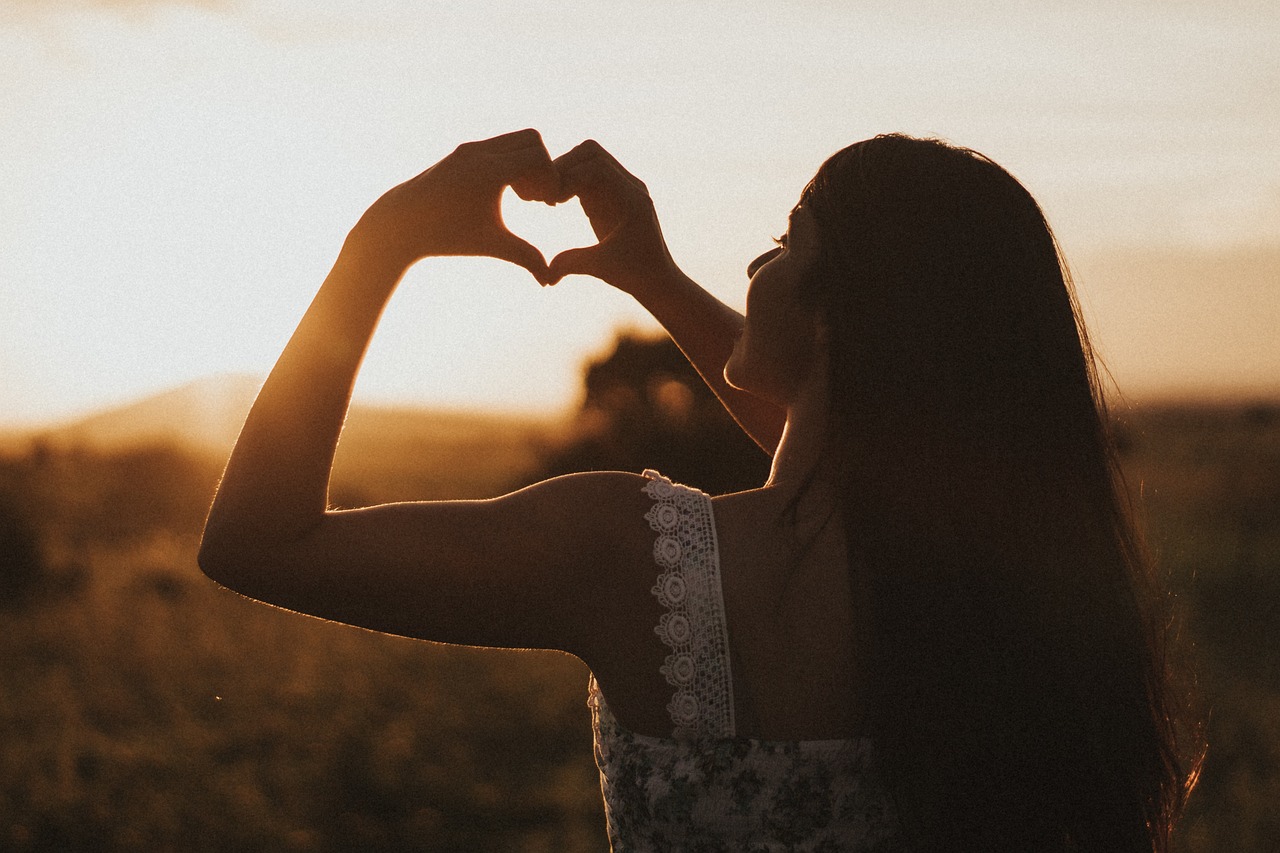 femme vue de dos, qui fait un cœur avec ses doigts, en arrière plan un coucher de soleil